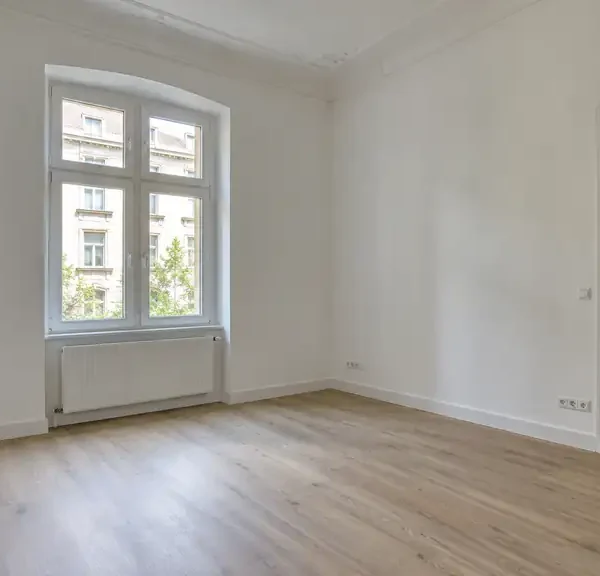 Frisch renovierter, leerer Raum in einer Altbauwohnung mit weißen Wänden, Stuck an der Decke, hellem Boden in Eichenoptik und hohem Sprossenfenster