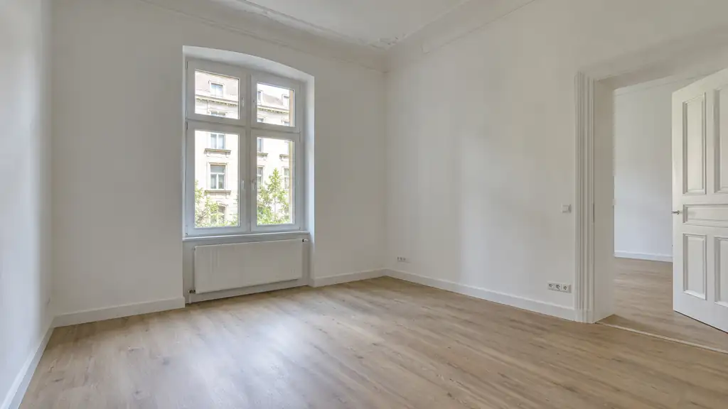 Frisch renovierter, leerer Raum in einer Altbauwohnung mit weißen Wänden, Stuck an der Decke, hellem Boden in Eichenoptik und hohem Sprossenfenster